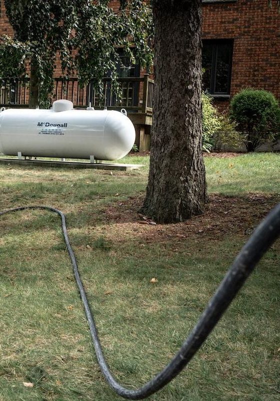 White propane tank installed outdoors on a grassy area near trees and a brick house.